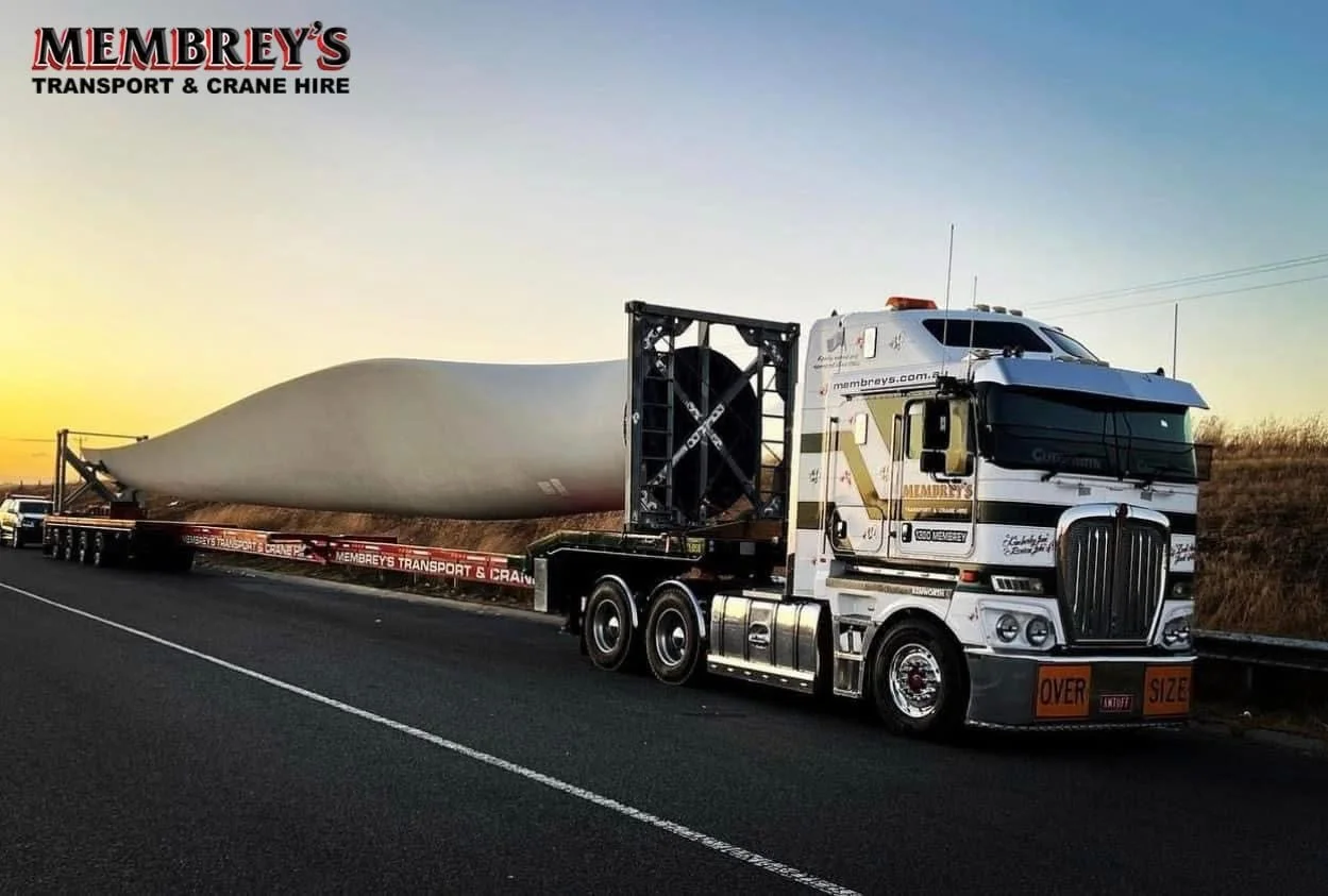 A large truck from Membrey's Transport & Crane Hire carries an oversized wind turbine blade on a trailer along a road at sunset.