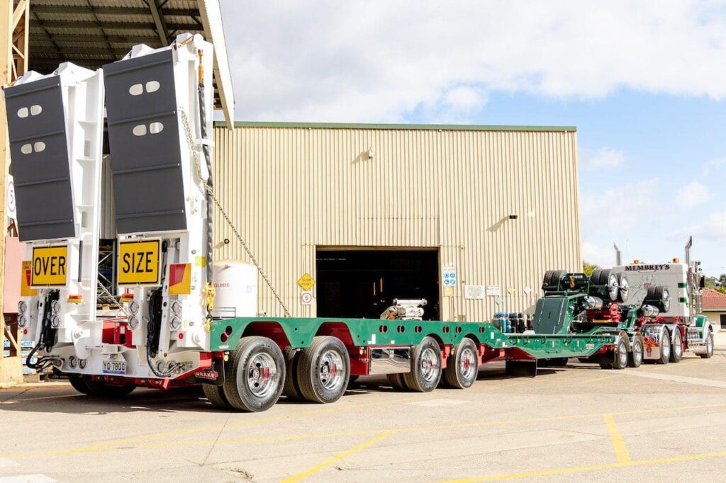 A large green and white flatbed trailer labeled "OVER SIZE" is parked outside a beige industrial building. Equipment is loaded on the trailer, and a semi-truck is attached at the front.