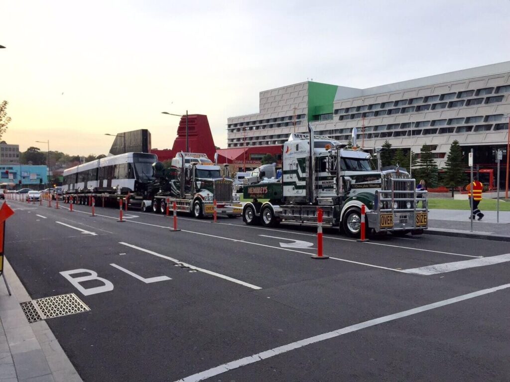 A large truck with a trailer is parked on a city street, carrying a train carriage. Orange safety cones line both sides of the truck, and modern buildings are visible in the background.