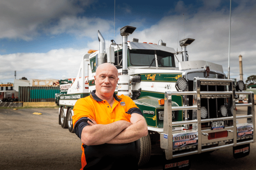A person in an orange and black shirt stands with arms crossed in front of a large green and white tow truck in an outdoor industrial area under a cloudy sky.