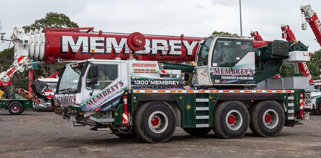 A large, multi-axle mobile crane with "Membrey's" branding on its cab and crane boom is parked in a lot, surrounded by other cranes and vehicles, with trees in the background.
