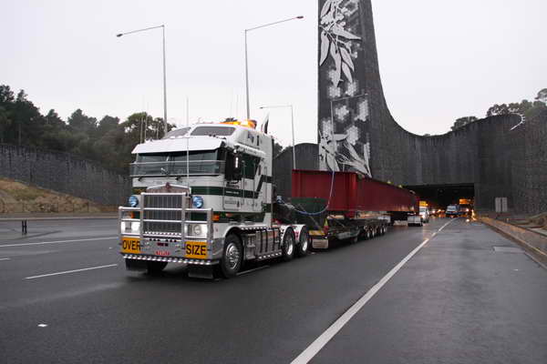 A large green and white "oversize" semi truck pulls a long red load on a trailer, driving on a multi-lane highway under a tall tunnel with decorative artwork. Other vehicles follow behind.