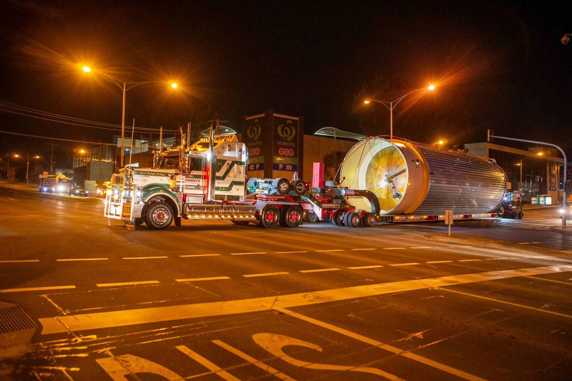 A large truck transports an oversized cylindrical structure through a city intersection at night, illuminated by streetlights and passing by a supermarket.