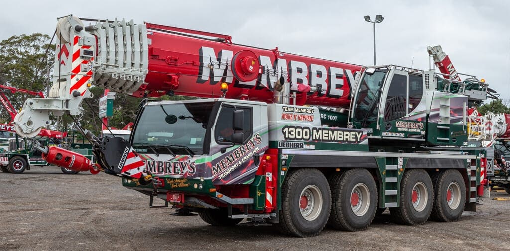 A large mobile crane truck with "Membrey" branding on the side and boom is parked in a lot, displaying its red and white crane arm and six large wheels.