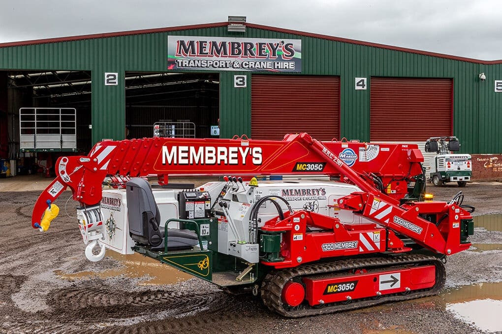 A red and white crane labeled "Membrey's" is parked outside a large green warehouse with "Membrey's Transport & Crane Hire" signage above the entrance on a cloudy day.