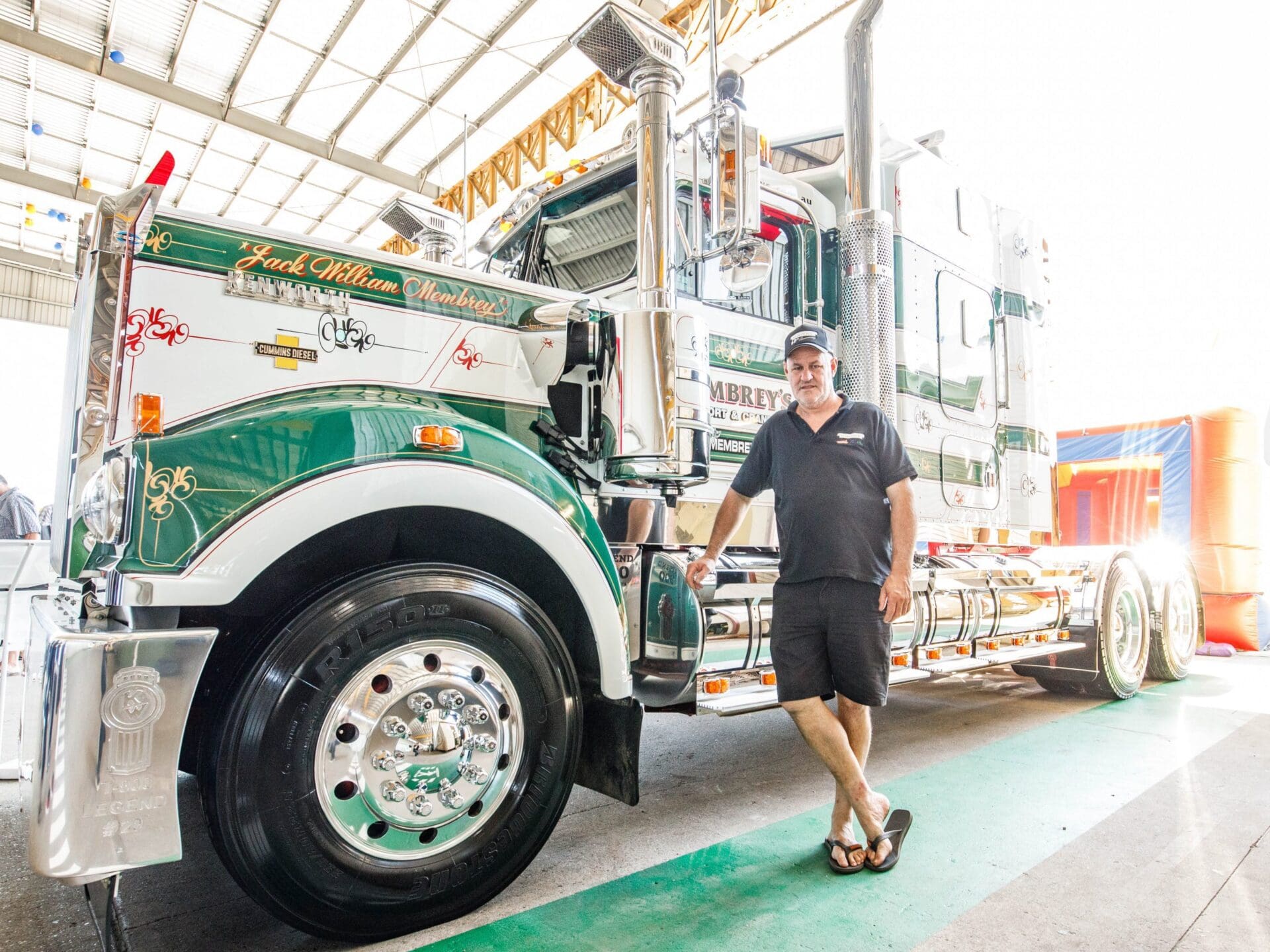 A man in a black shirt, shorts, and cap stands smiling next to a large, green and white semi-truck inside a well-lit garage or exhibition space.