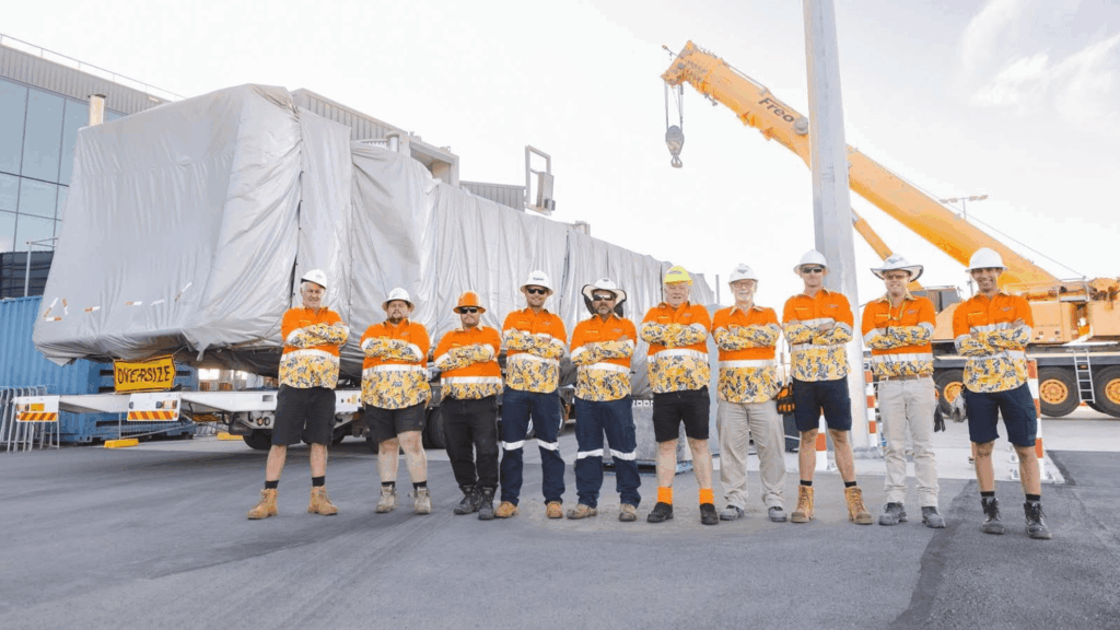 A group of ten construction workers in orange and yellow high-visibility shirts stand in front of a large, covered piece of equipment being transported by a crane and truck on a construction site.