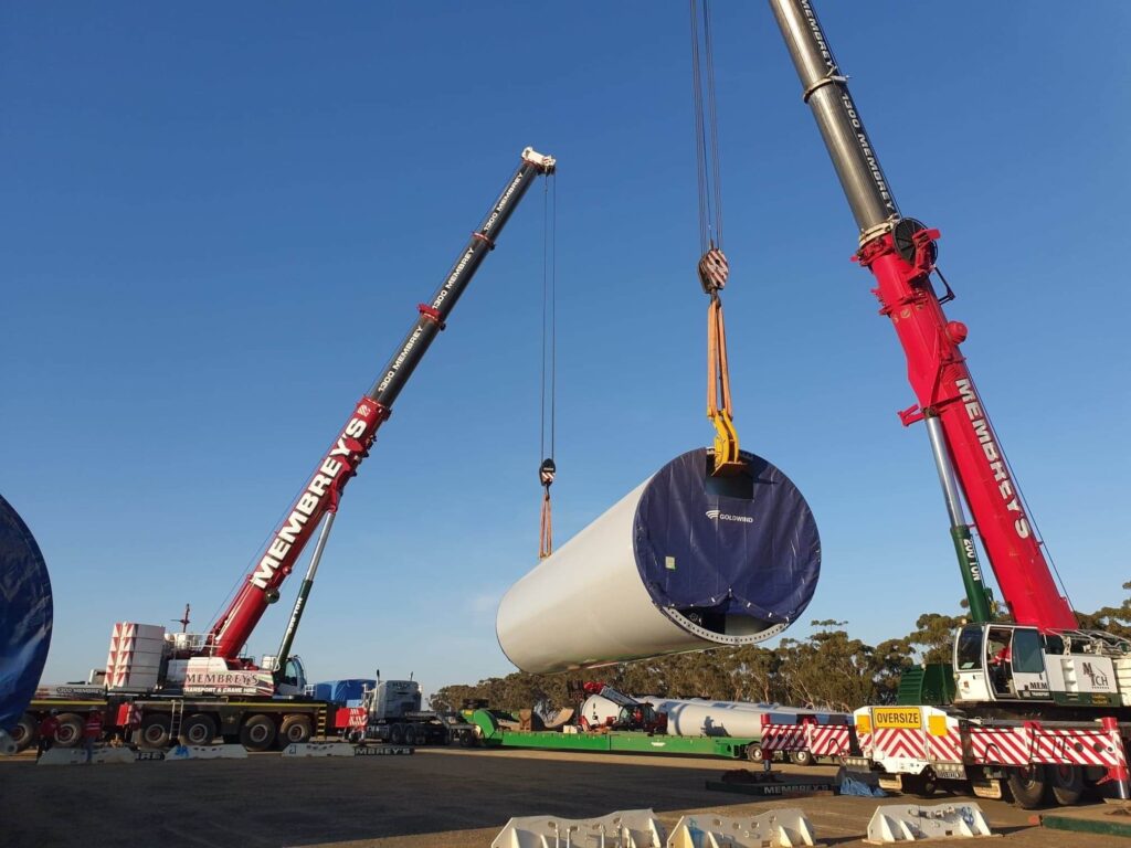 Two large cranes lift a cylindrical wind turbine tower section at a construction site, with trees and blue sky in the background. The cranes and equipment are branded with company names.