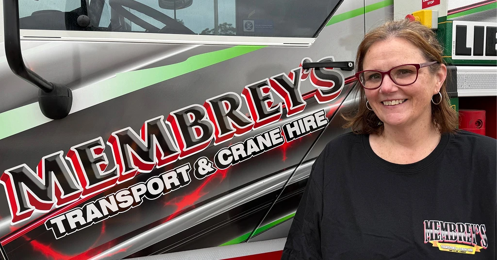A woman with glasses and brown hair smiles while standing next to a truck with "Membrey’s Transport & Crane Hire" written on the door. She is wearing a black shirt with the same logo.