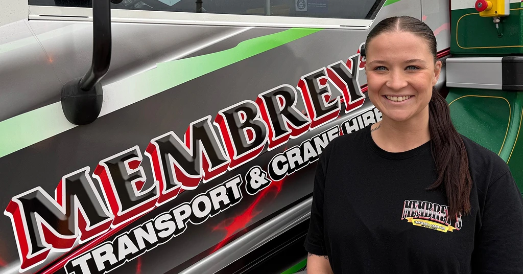 A smiling woman with dark hair tied back stands beside a truck displaying the logo "Membrey's Transport & Crane Hire" in bold red and white lettering. She wears a black Membrey's t-shirt.