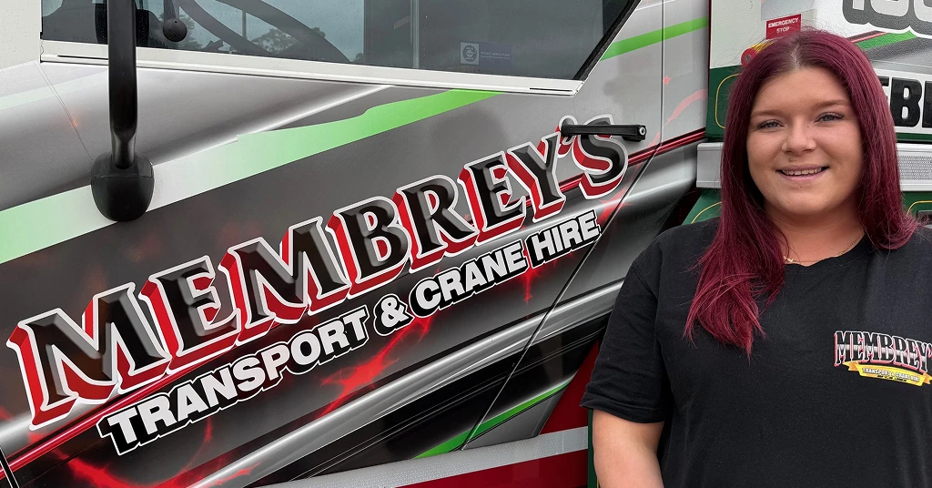A woman with long reddish hair stands smiling next to a truck with the logo “Membrey’s Transport & Crane Hire” on the side. She wears a black Membrey’s t-shirt.